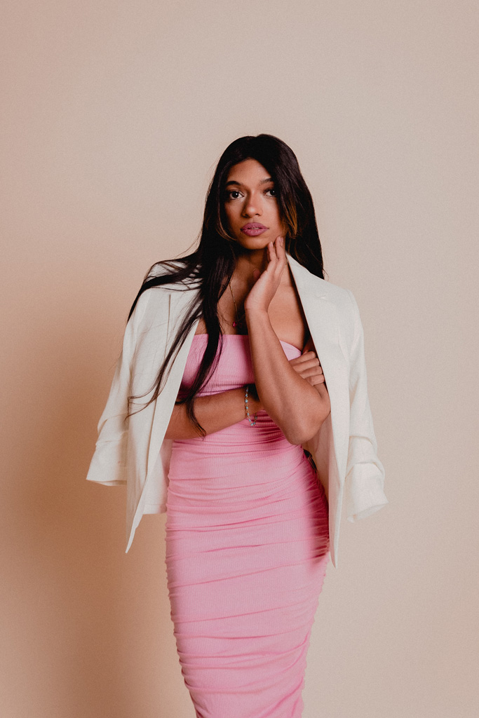 Coach is posing in an elegant pink dress and a white blazer with her hair down.